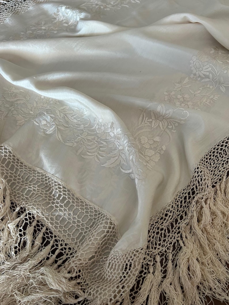 K&ouml;nnte beinhalten: Cremefarbener Schal mit floraler Stickerei und geh&auml;kelter Fransenborte. Der Stoff ist in weichen Falten drapiert, wodurch die filigranen Details hervorgehoben werden. Die Fransen verleihen den Schalkanten Textur.