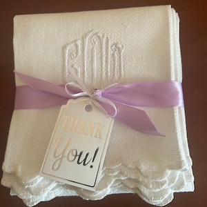 May include: A stack of white, scalloped-edge towels tied with a lavender ribbon and a silver tag that reads "Thank You!". The towels have an embroidered monogram design.