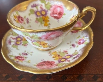 May include: A porcelain teacup and saucer set featuring a white base with a floral pattern of pink roses, yellow blossoms, and purple flowers. The edges of both the cup and saucer are scalloped and trimmed with a metallic gold painted finish.