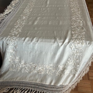K&ouml;nnte beinhalten: Cremefarbene rechteckige Tischdecke mit floraler Stickerei. Die R&auml;nder haben einen geh&auml;kelten Rand und Fransen. Die gestickten Blumenmuster verleihen dem Textil Eleganz, passend f&uuml;r einen Esstisch.