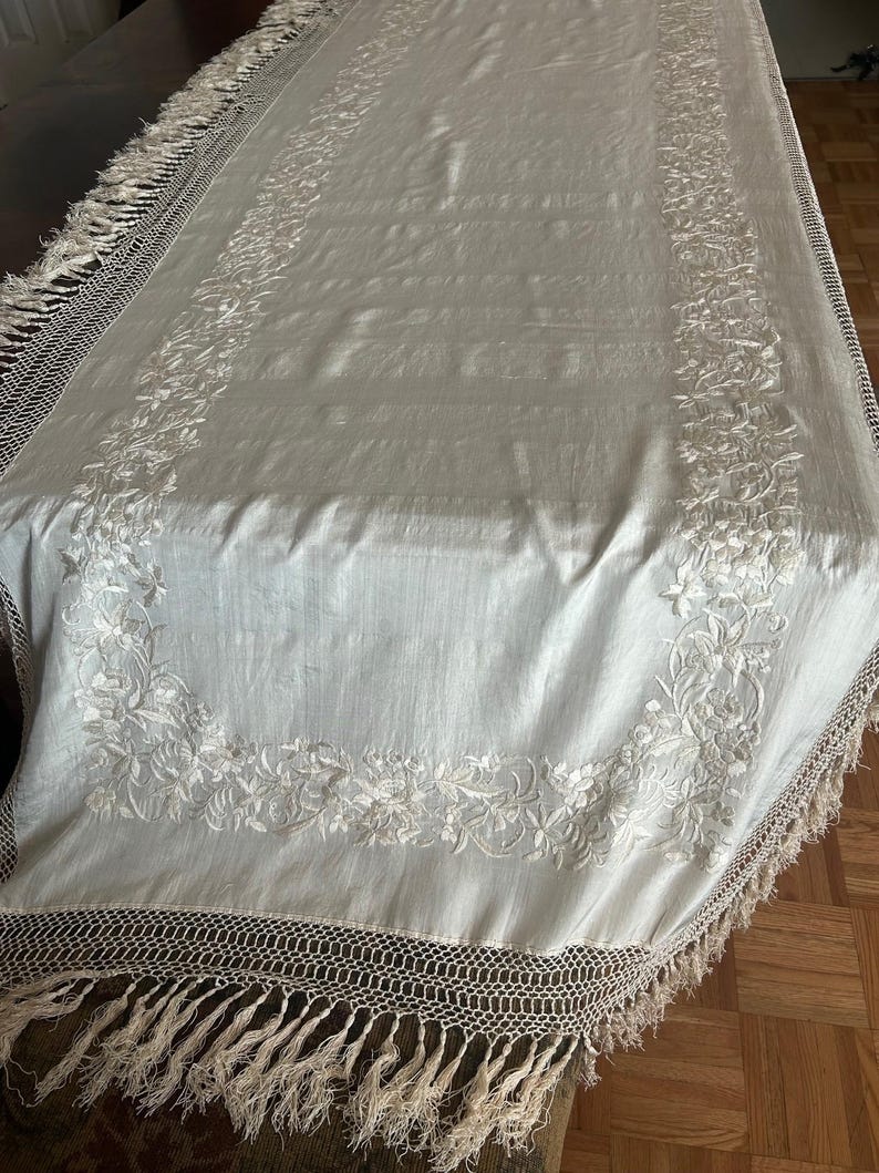 K&ouml;nnte beinhalten: Cremefarbene rechteckige Tischdecke mit floraler Stickerei. Die R&auml;nder haben einen geh&auml;kelten Rand und Fransen. Die gestickten Blumenmuster verleihen dem Textil Eleganz, passend f&uuml;r einen Esstisch.