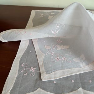 May include: White linen placemat with a scalloped edge and delicate pink floral embroidery. The placemat is folded, revealing the intricate design and texture. A classic and elegant piece for dining.