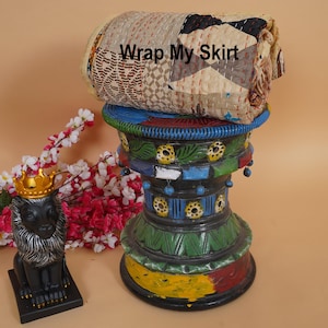Könnte beinhalten: Gerolltes Patchwork-Textil mit dem Text "Wrap My Skirt" auf einem farbenfrohen, geschnitzten Sockel. Das Textil weist verschiedene geometrische Muster in neutralen Tönen auf. Eine schwarze Löwenstatue mit goldener Krone befindet sich links.