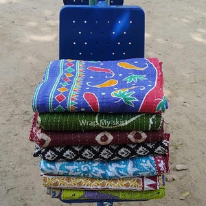 May include: A stack of colorful kantha quilts with various patterns, including floral, geometric, and paisley designs. The quilts are folded and stacked on a blue metal chair. The text "Wrap My Skirt" is visible on the green quilt.