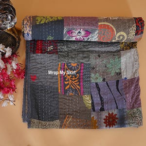 May include: A patchwork quilt with a variety of muted colors, including gray, purple, and green. The quilt is made of different fabric squares stitched together with visible white thread. The text "Wrap My Skirt" is visible on the quilt.