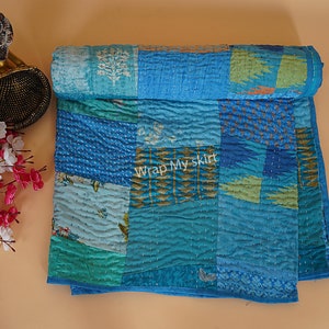 May include: A blue and green patchwork quilt with a kantha stitch pattern. The quilt is made of recycled saris and is hand-stitched. The quilt is folded in half and is lying on a light brown surface. The text "Wrap My Skirt" is visible in the center of the quilt.