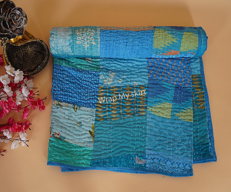 May include: A blue and green patchwork quilt with a kantha stitch pattern. The quilt is made of recycled saris and is hand-stitched. The quilt is folded in half and is lying on a light brown surface. The text "Wrap My Skirt" is visible in the center of the quilt.