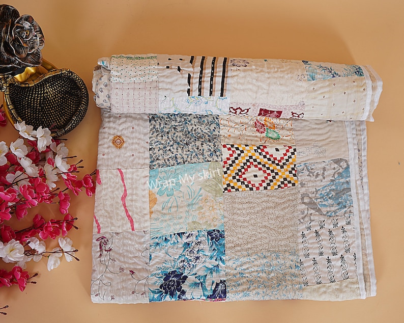 May include: A white kantha quilt with a patchwork design featuring various patterns and colors, including blue, red, and yellow. The quilt is made of recycled saris and is hand-stitched. "Wrap My Skirt" is written on the quilt.