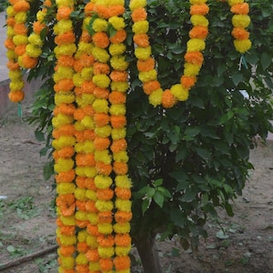 Könnte beinhalten: Girlanden aus gelben und orangen Ringelblumen sind über einen grünen Busch drapiert. Die Girlanden sind dicht mit runden Blüten gepackt und erzeugen eine lebendige und festliche Darstellung. Das Bild zeigt die leuchtenden Farben der Blumen.