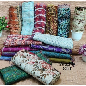 May include: A collection of colorful kantha quilts, also known as kantha stitch quilts, made from recycled saris. The quilts are folded and stacked on a light brown surface. The text "WrapMySkirt" is visible in the lower right corner of the image.