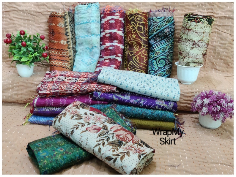 May include: A collection of colorful kantha quilts, also known as kantha stitch quilts, made from recycled saris. The quilts are folded and stacked on a light brown surface. The text "WrapMySkirt" is visible in the lower right corner of the image.