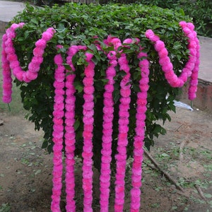May include: A garland of pink fabric flowers, arranged in a vertical line, hanging from a bush. The garland is made of multiple strands of flowers, each strand is about 90 cm long.