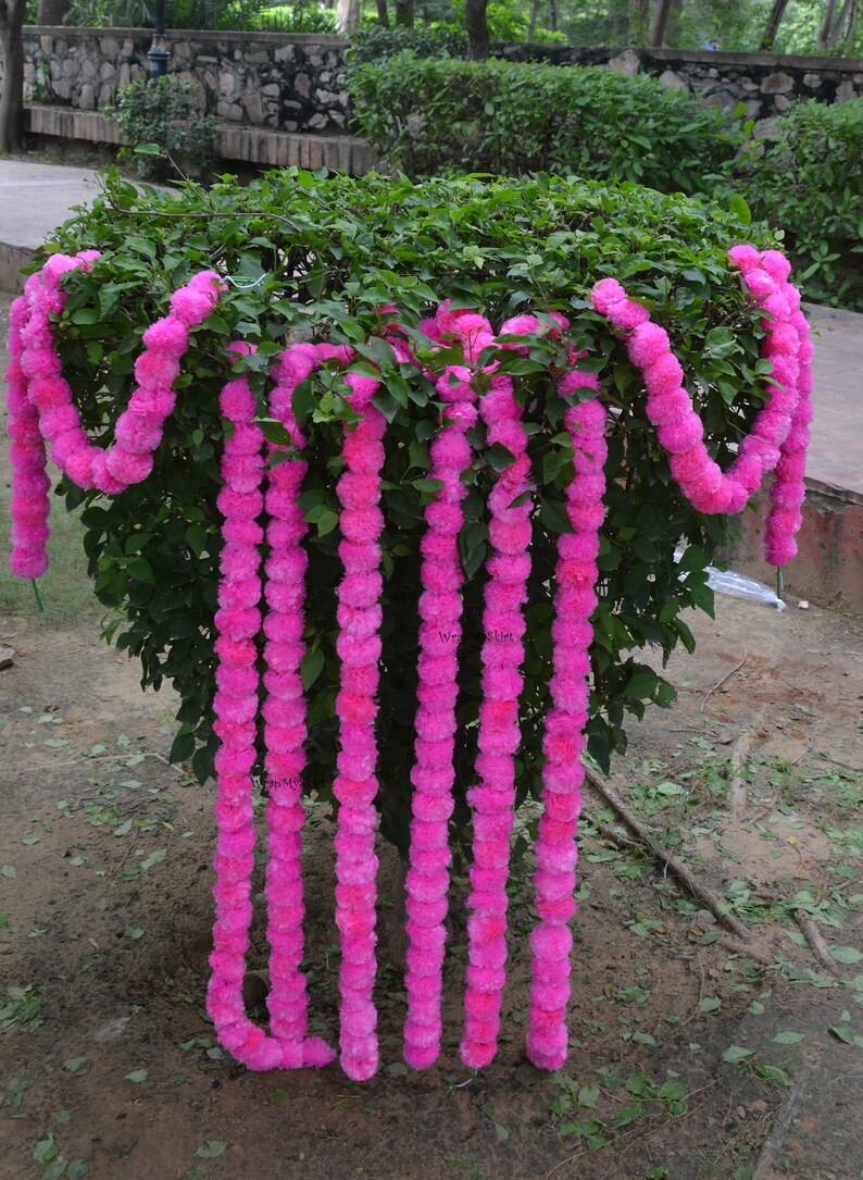 May include: A garland of pink fabric flowers, arranged in a vertical line, hanging from a bush. The garland is made of multiple strands of flowers, each strand is about 90 cm long.