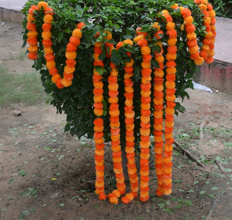 May include: A set of 8 orange and yellow marigold flower garlands hanging from a tree branch. The garlands are made of artificial flowers and are perfect for decorating for a wedding or other special occasion.