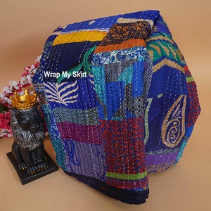 Könnte beinhalten: Eine gefaltete Patchwork-Decke mit verschiedenen blauen, gelben und orangefarbenen Stoffbahnen mit komplizierten Mustern und weißen Kantha-Nähten. Der Text Wrap My Skirt ist auf dem Stoff sichtbar. Eine schwarze Löwenfigur mit goldener Krone.