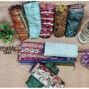 May include: A collection of colourful kantha quilts, also known as kantha stitch quilts, in various patterns and colours. The quilts are rolled up and arranged on a light brown surface. The text "WrapMySkirt" is visible in the image.