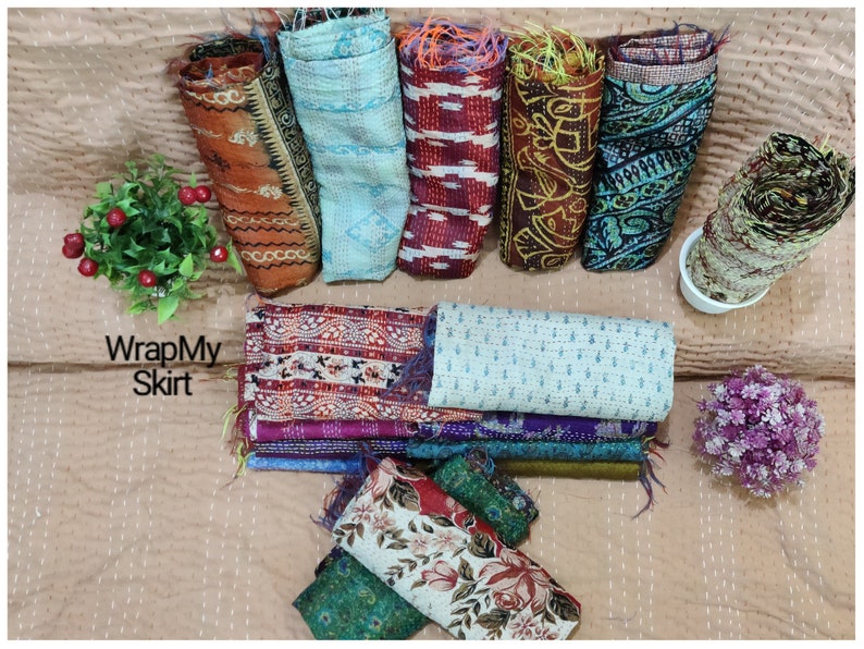 May include: A collection of colourful kantha quilts, also known as kantha stitch quilts, in various patterns and colours. The quilts are rolled up and arranged on a light brown surface. The text "WrapMySkirt" is visible in the image.