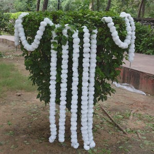 Könnte beinhalten: Weiße Blumengirlanden, die über einem grünen Busch drapiert sind. Die Girlanden bestehen aus runden, flauschigen weißen Blüten und hängen in vertikalen Strängen. Der Hintergrund umfasst grünes Laub und einen Blick auf einen Weg.