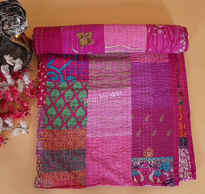 May include: A pink kantha quilt with a patchwork design featuring various floral and geometric patterns. The quilt is made of recycled saris and is hand-stitched.