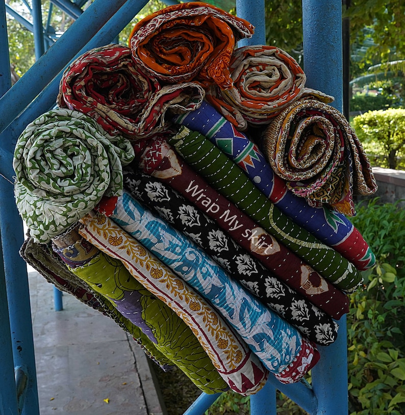 May include: A stack of colourful, hand-stitched kantha quilts, rolled up and tied together. The quilts are made from recycled saris and feature intricate patterns and designs. The text "Wrap My Skirt" is visible on the quilts.