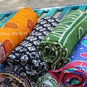 May include: A collection of colourful, rolled up kantha quilts with various patterns, including paisley, floral, and geometric designs. The quilts are stacked on a green metal rack. The text "Wrap My Skirt" is visible on the orange quilt.