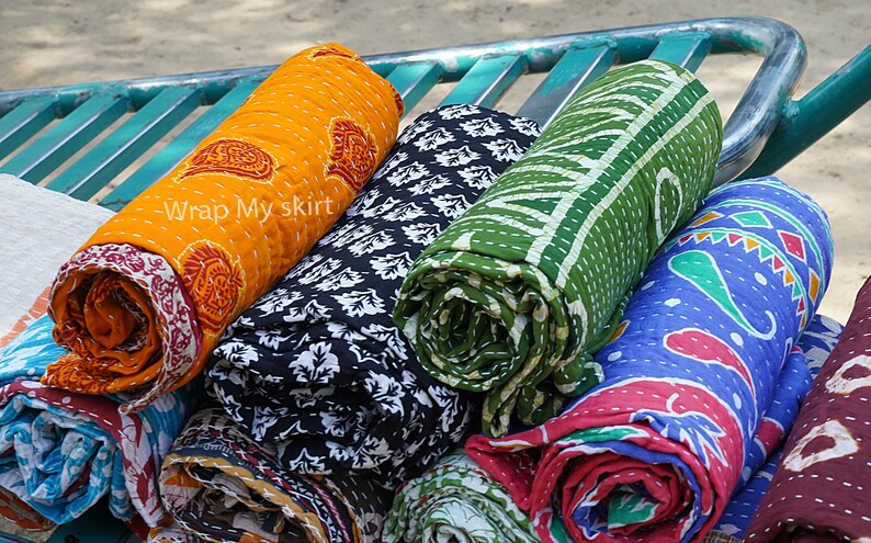 May include: A collection of colourful, rolled up kantha quilts with various patterns, including paisley, floral, and geometric designs. The quilts are stacked on a green metal rack. The text "Wrap My Skirt" is visible on the orange quilt.
