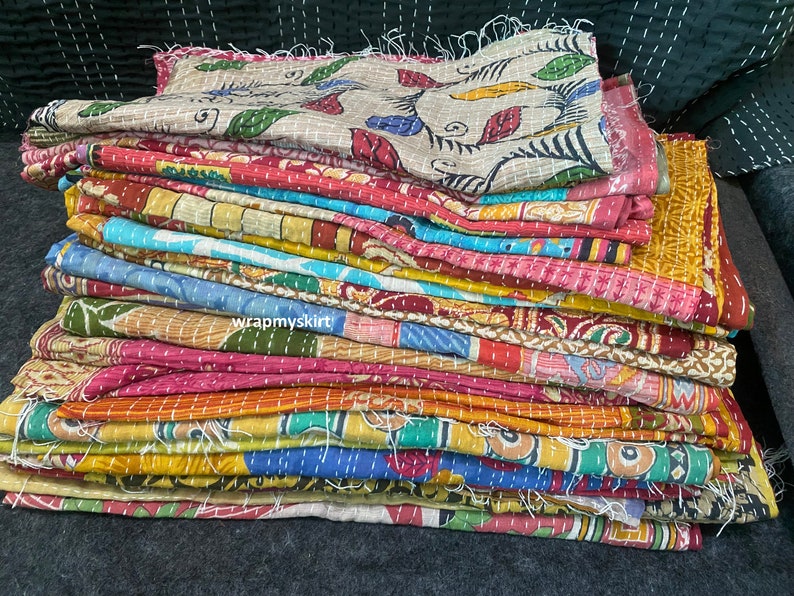 Op de afbeelding: Een stapel kleurrijke kantha-dekens met verschillende bloemen- en geometrische patronen. De dekens zijn gemaakt van gerecyclede sari's en zijn met de hand gestikt. De dekens zijn gevouwen en gestapeld op een donker oppervlak. De tekst "wrapmyskirts" is zichtbaar aan de onderkant van de stapel.