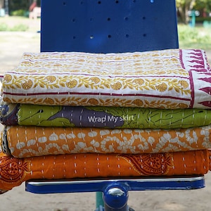 May include: Four kantha quilts stacked on a blue chair. The top quilt is white with a yellow floral pattern and a burgundy border. The second quilt is green with a purple and white floral pattern. The third quilt is orange with a white polka dot pattern. The bottom quilt is orange with a red paisley pattern.