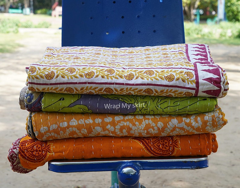 May include: Four kantha quilts stacked on a blue chair. The top quilt is white with a yellow floral pattern and a burgundy border. The second quilt is green with a purple and white floral pattern. The third quilt is orange with a white polka dot pattern. The bottom quilt is orange with a red paisley pattern.