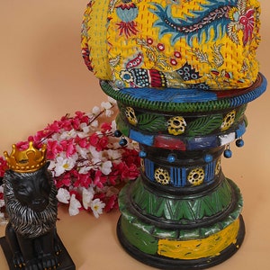 May include: A yellow, floral patterned kantha quilt is rolled up and placed on a colorful, hand-painted wooden stand. A black lion figurine with a gold crown sits on a black base in the foreground.