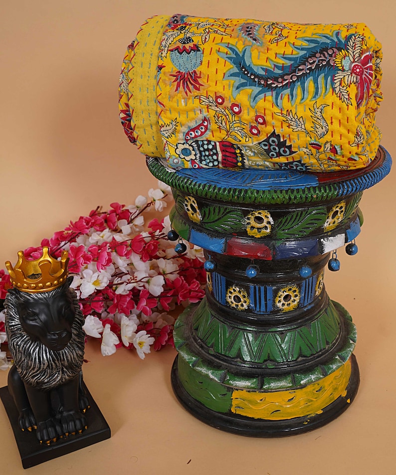May include: A yellow, floral patterned kantha quilt is rolled up and placed on a colorful, hand-painted wooden stand. A black lion figurine with a gold crown sits on a black base in the foreground.