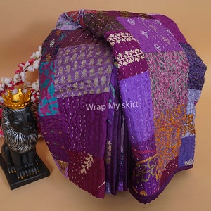 Puede incluir: Una colcha de retazos morada y dorada hecha de tela de seda con puntadas intrincadas. La colcha est&aacute; doblada y dispuesta sobre una superficie marr&oacute;n claro. El texto "Wrap My Skirt" es visible en la colcha.