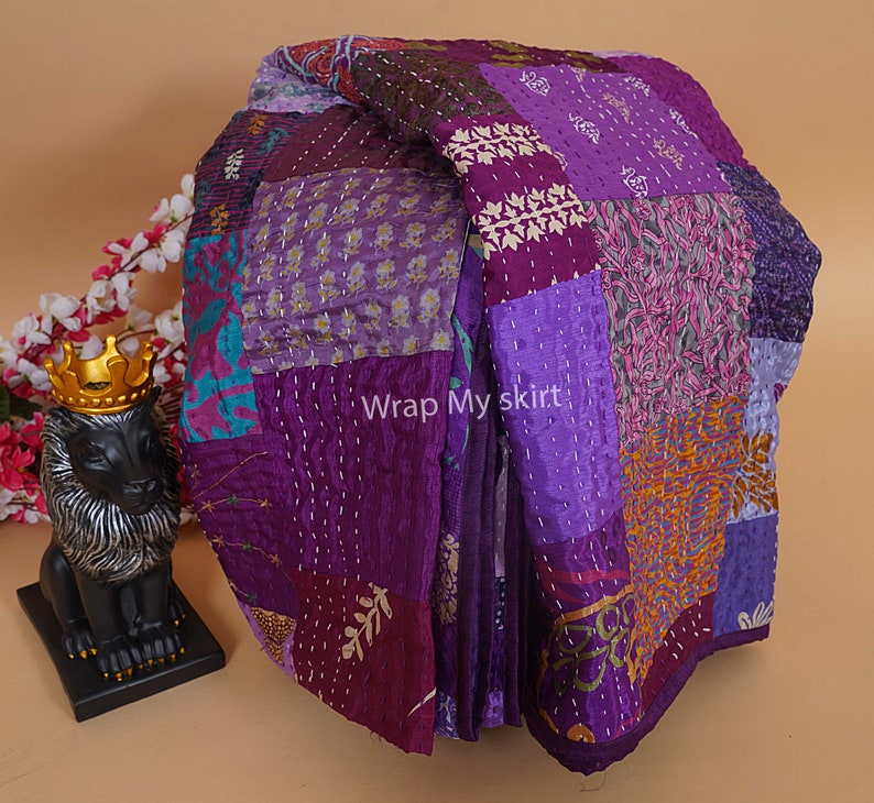 Puede incluir: Una colcha de retazos morada y dorada hecha de tela de seda con puntadas intrincadas. La colcha est&aacute; doblada y dispuesta sobre una superficie marr&oacute;n claro. El texto "Wrap My Skirt" es visible en la colcha.