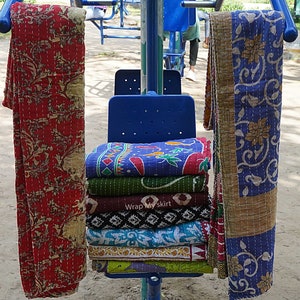 Könnte beinhalten: Ein Stapel bunter Kantha-Steppdecken, ein traditionelles indisches Textil, wird auf einem blauen Metallstuhl ausgestellt. Die Decken sind gefaltet und gestapelt, wobei die oberste Decke ein leuchtendes Lila mit einem Blumenmuster hat. Die Decken hängen an einem Metallgestell, wobei der Text "Wrap My Skirt" auf dem Stuhl sichtbar ist.