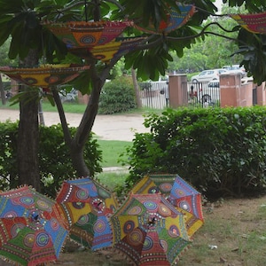 Könnte beinhalten: Eine Sammlung farbenfroher, dekorativer Schirme mit aufwendigen Mustern, einige hängen an einem Baum, andere liegen auf dem Boden. Die Schirme weisen eine Vielzahl von Farben auf, darunter Rot, Blau, Gelb und Grün, was ein lebendiges Bild ergibt.