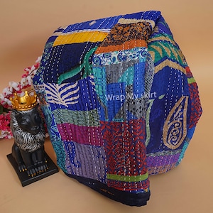 Pu&ograve; includere: Una trapunta kantha blu e multicolore con un design a patchwork. La trapunta &egrave; realizzata con sari riciclati e presenta una variet&agrave; di motivi floreali e geometrici. La trapunta &egrave; piegata e appoggiata su una superficie marrone chiaro. "Wrap My Skirt" &egrave; scritto in bianco sulla trapunta.