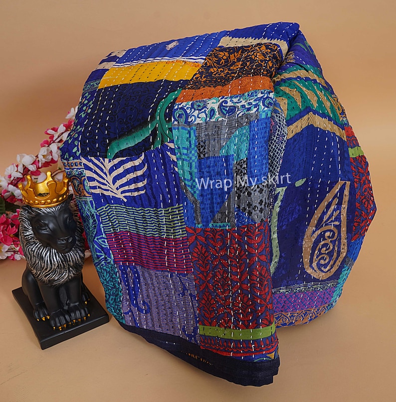 Pu&ograve; includere: Una trapunta kantha blu e multicolore con un design a patchwork. La trapunta &egrave; realizzata con sari riciclati e presenta una variet&agrave; di motivi floreali e geometrici. La trapunta &egrave; piegata e appoggiata su una superficie marrone chiaro. "Wrap My Skirt" &egrave; scritto in bianco sulla trapunta.