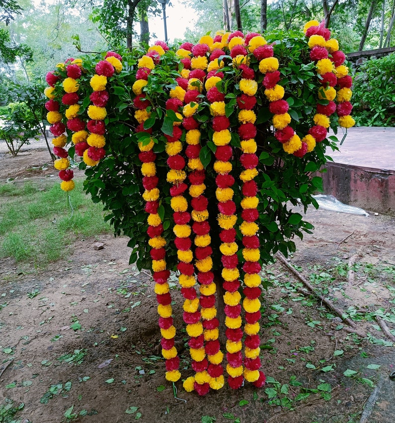 May include: A garland of yellow and red artificial flowers, strung together in a long chain, hangs from a tree branch. The garland is made of fabric flowers and is a popular decoration for weddings and other special events.