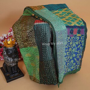 Pu&ograve; includere: Una trapunta patchwork realizzata con diversi tessuti in tonalit&agrave; di verde, blu e giallo. La trapunta &egrave; cucita a mano con filo bianco e ha un bordo sfrangiato. La trapunta &egrave; piegata ed esposta su una superficie marrone. "Wrap My Skirt" &egrave; stampato sulla trapunta.
