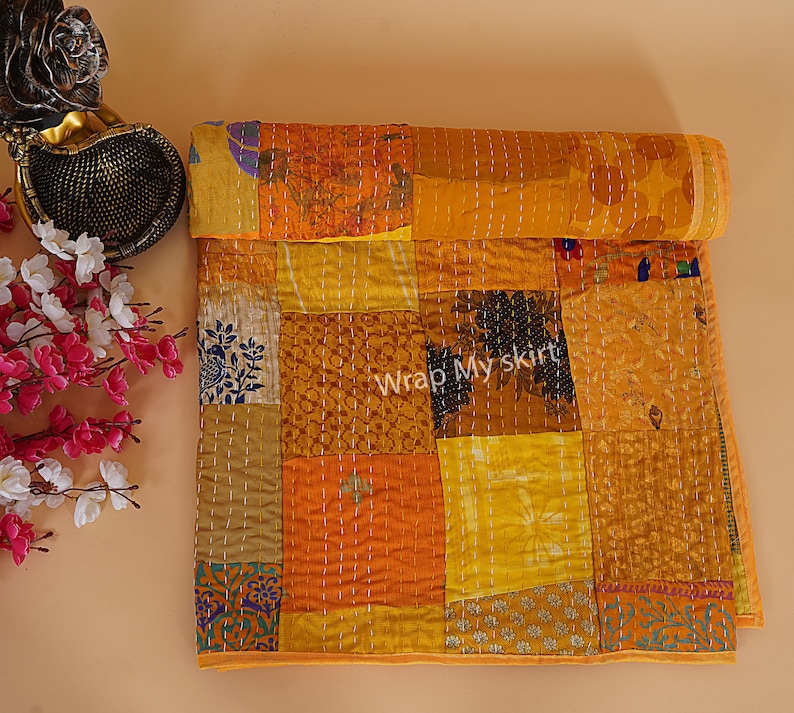 May include: A yellow and orange patchwork quilt with a variety of patterns and textures. The quilt is made of recycled saris and is hand-stitched. The quilt is rolled up and has a tag that says "Wrap My Skirt".