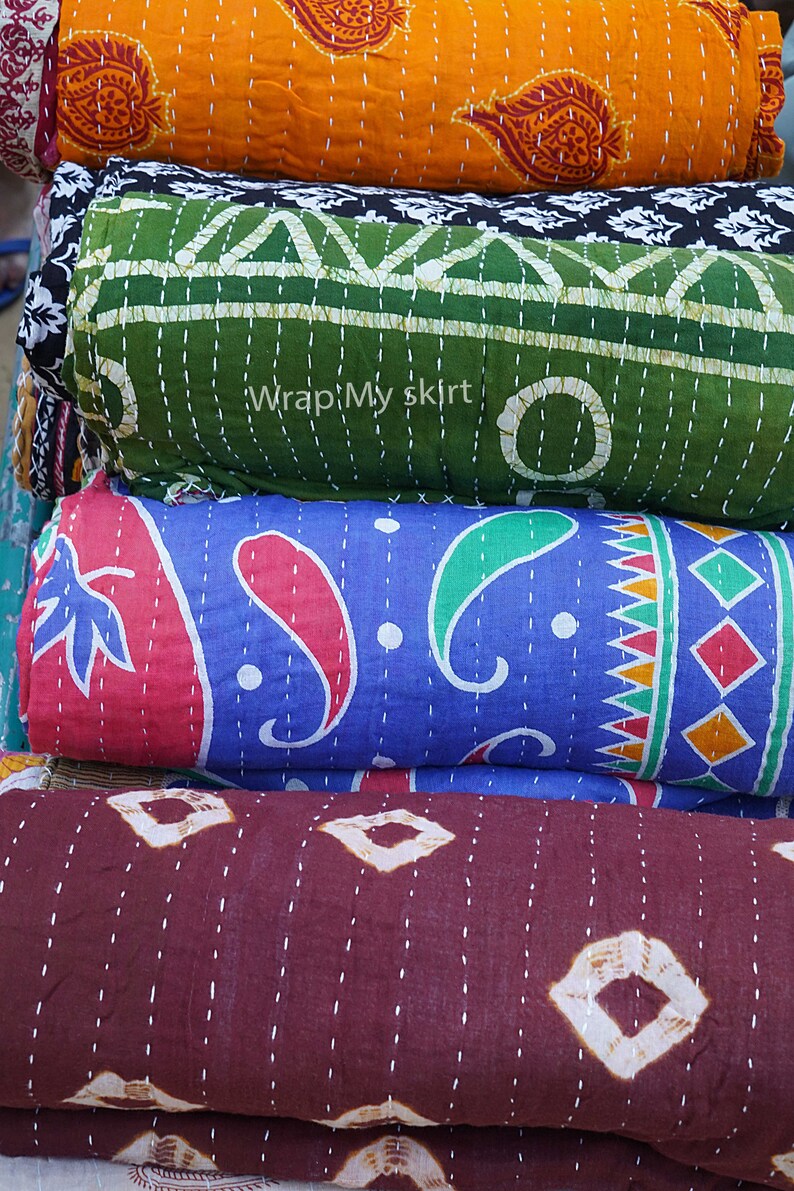 May include: A stack of four kantha quilts in various colours and patterns. The top quilt is orange with a floral design, the second is green with a geometric design, the third is blue with a paisley design, and the bottom quilt is brown with a diamond pattern. The quilts are all hand-stitched and have a vintage look.