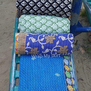 Könnte beinhalten: Ein Stapel bunter Kantha-Steppdecken mit floralen und geometrischen Mustern. Die Decken sind gefaltet und auf einem Metallgestell angeordnet. Der Text "Wrap My Skirt" ist auf der untersten Decke sichtbar.