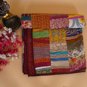 May include: A colorful patchwork quilt made from recycled saris. The quilt is made up of squares of different colors and patterns, including red, orange, yellow, green, blue, and purple. The quilt is folded in half and has a maroon border.