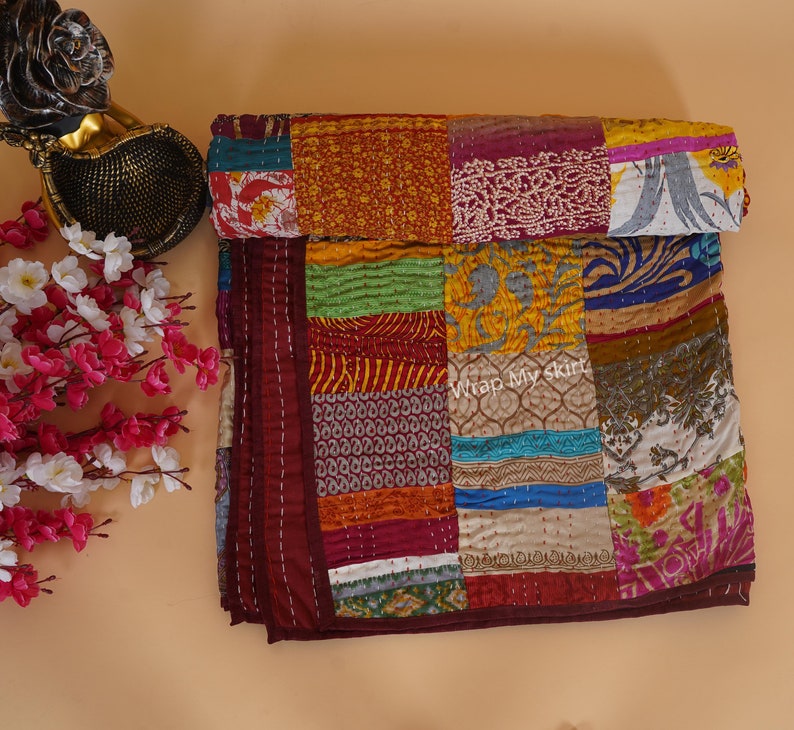 May include: A colorful patchwork quilt made from recycled saris. The quilt is made up of squares of different colors and patterns, including red, orange, yellow, green, blue, and purple. The quilt is folded in half and has a maroon border.