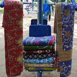 May include: A stack of colourful kantha quilts, folded and displayed on a blue chair. The quilts are made of cotton and have a variety of patterns, including floral and geometric designs. The quilts are a traditional Indian craft and are known for their durability and beauty.