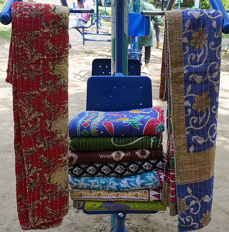 May include: A stack of colourful kantha quilts, folded and displayed on a blue chair. The quilts are made of cotton and have a variety of patterns, including floral and geometric designs. The quilts are a traditional Indian craft and are known for their durability and beauty.