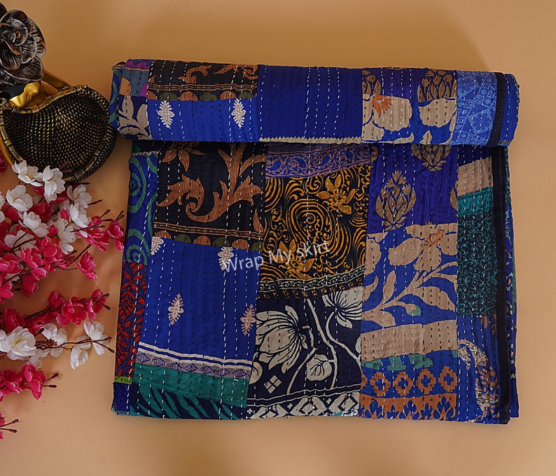 May include: A patchwork quilt with a blue background and various floral and geometric patterns in shades of brown, gold, green, and white. The quilt is made of recycled saris and is hand-stitched.