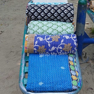May include: Four colourful kantha quilts rolled up and stacked on a metal rack. The quilts have floral and geometric patterns in shades of blue, green, black, and white. The text "Wrap My Skirt" is visible on the bottom quilt.