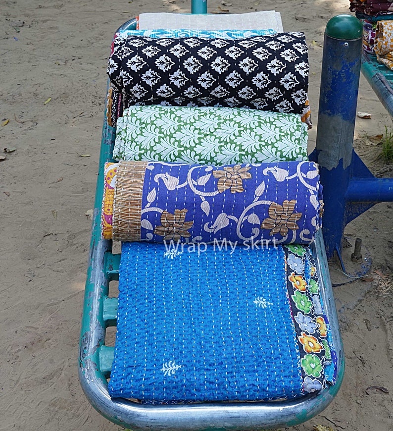 May include: Four colourful kantha quilts rolled up and stacked on a metal rack. The quilts have floral and geometric patterns in shades of blue, green, black, and white. The text "Wrap My Skirt" is visible on the bottom quilt.