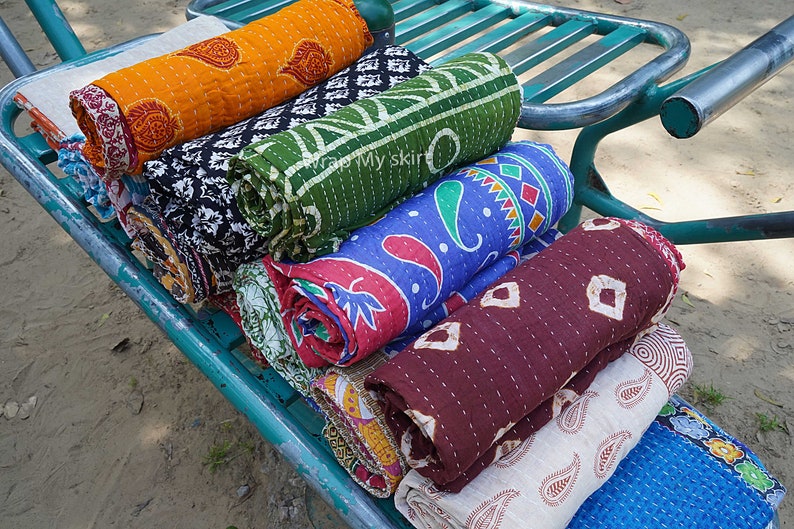 May include: A stack of colourful kantha quilts rolled up and displayed on a metal cart. The quilts are made of cotton and have intricate patterns in shades of blue, green, red, orange, and brown.